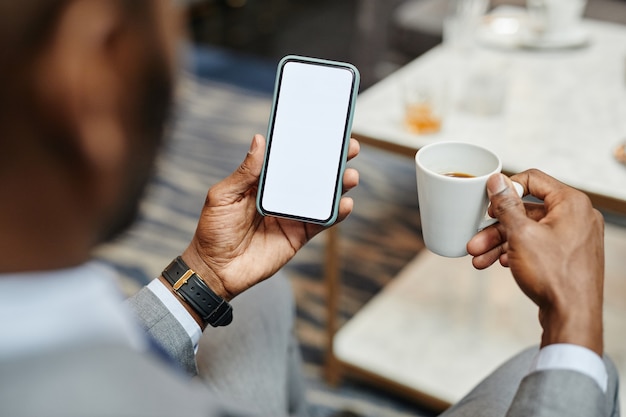 Nahaufnahme eines erfolgreichen afroamerikanischen Geschäftsmannes, der Smartphone mit leerem Bildschirm hält, während er Kaffee im Café genießt, Kopienraum