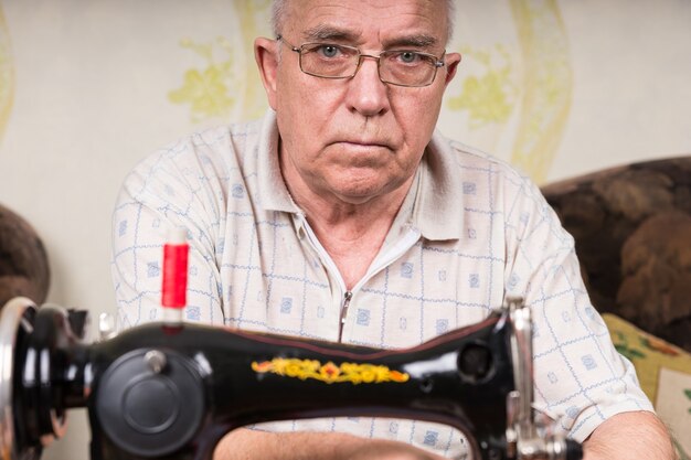 Nahaufnahme eines ernsthaften älteren Mannes mit Brille mit altmodischer Nähmaschine zu Hause im Wohnzimmer