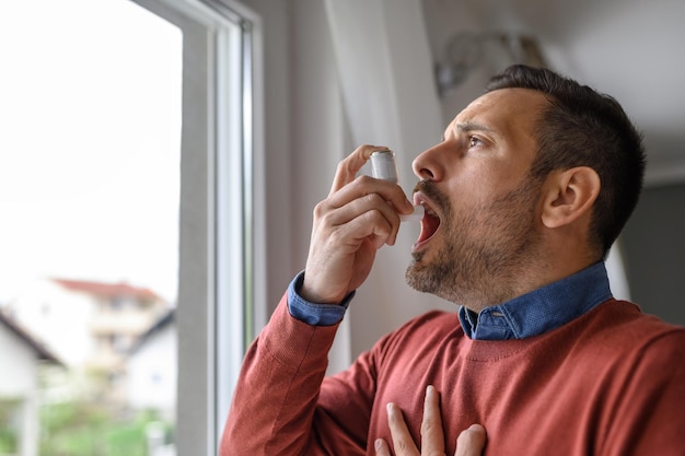 Nahaufnahme eines erstickten jungen Mannes, der zu Hause durch eine Asthmapumpe am Fenster einatmet