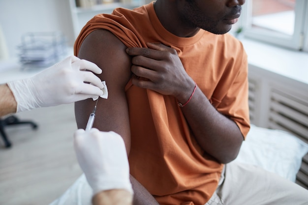 Nahaufnahme eines erwachsenen afroamerikanischen Mannes, der wegschaut, während er in einer Klinik oder einem Krankenhaus einen Covid-Impfstoff erhält, wobei eine männliche Krankenschwester den Impfstoff in die Schulter injiziert