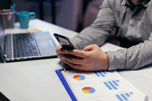 Nahaufnahme eines Freiberuflers, der Überstunden auf dem Smartphone-Sitzarbeitsplatz schreibt. Geschäftsmann, der seine Zelle verwendet, um eine SMS zu senden, während er spät nachts im Büro arbeitet, um eine Frist zu beenden.