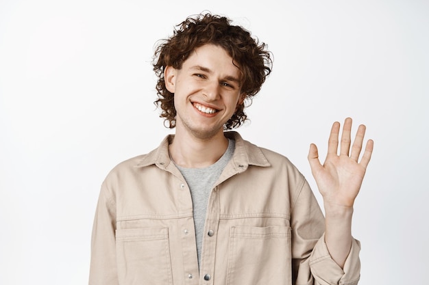 Foto nahaufnahme eines freundlich lächelnden jungen mannes winkt mit der hand und lächelt, um sie zu begrüßen und hallo zu sagen, steht auf weißem hintergrund