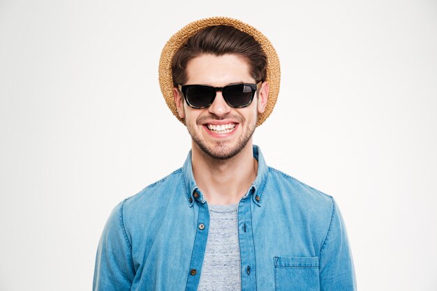 Nahaufnahme eines fröhlichen jungen Mannes mit Hut und Sonnenbrille, der auf weißem Hintergrund steht und lächelt