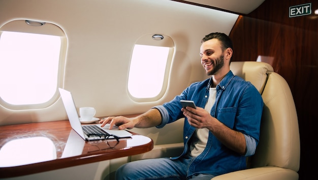 Nahaufnahme eines fröhlichen Mannes in einem lässigen Outfit, der seinen Flug genießt, auf seinem Fensterplatz sitzt und Nachrichten auf seinem Smartphone austauscht.