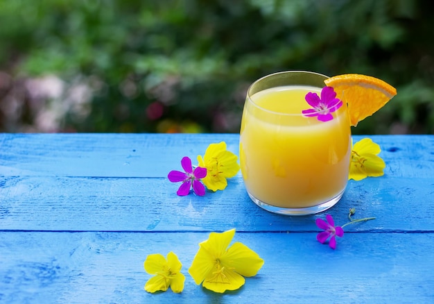 Nahaufnahme eines Glases frisch gepresster Orangensaft, garniert mit einer Scheibe reifer Orange und Blumen auf dem blauen Holztisch