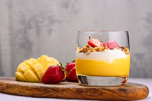 Nahaufnahme eines Glases mit einem fruchtigen Dessert und einem Löffel aus der Mango, Erdbeeren, Müsli und Ricotta auf Holz