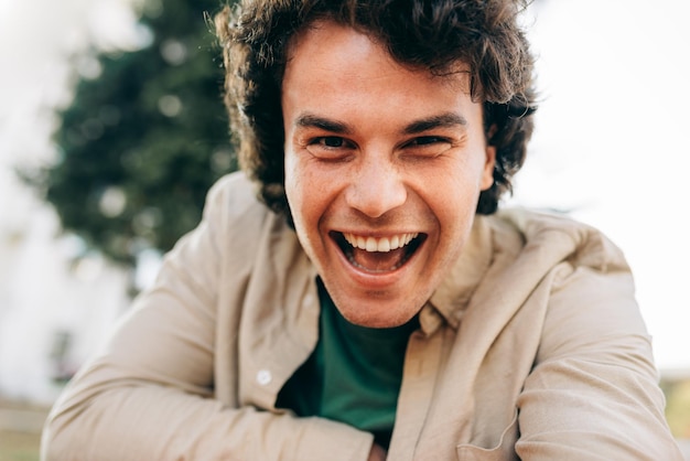 Nahaufnahme eines glücklichen jungen Mannes, der breit mit gesunden, zahnigen Zähnen lächelt und in die Kamera schaut Außenporträt eines glücklichen, intelligenten Studenten, der draußen ruht Menschen- und Emotionskonzept