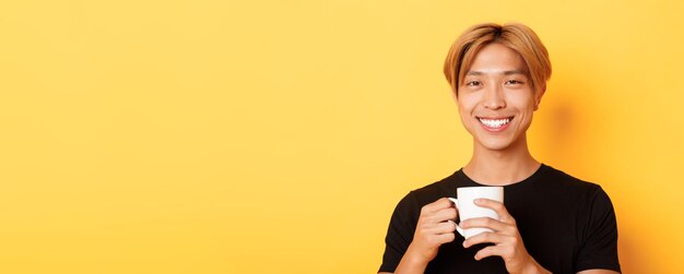 Nahaufnahme eines glücklichen, zufriedenen asiatischen lächelnden Kerls, der einen Becher mit Kaffeetrinken hält und zufrieden aussieht st