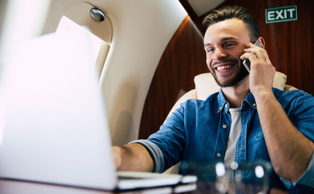 Nahaufnahme eines gut aussehenden Mannes in einem lässigen Outfit, der lächelt, während er etwas auf seinem Laptop tippt und ein Smartphone in der linken Hand hält und ein Flugzeug der ersten Klasse fliegt.