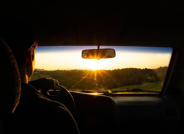 Nahaufnahme eines hispanischen jungen Mannes, der ein Auto fährt, während die Sonne am Horizont aufgeht