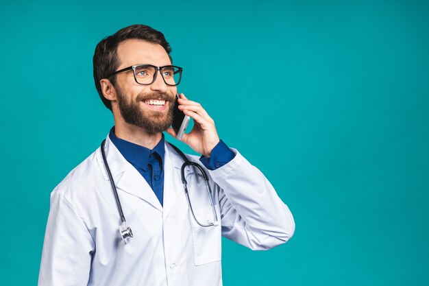 Nahaufnahme eines hübschen jungen Arztes auf blauem Hintergrund, der auf dem Smartphone spricht und positiv lächelt. Handy benutzen.