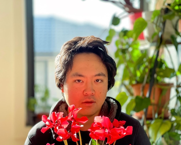Foto nahaufnahme eines jungen asiatischen mannes, der eine rot blühende zyklamenpflanze gegen hauspflanzen und ein fenster hält