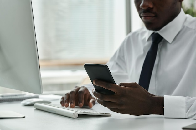 Nahaufnahme eines jungen Geschäftsmannes, der sein Smartphone benutzt, während er am Tisch im Büro am Computer arbeitet