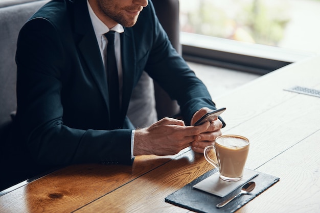 Nahaufnahme eines jungen Geschäftsmannes im vollen Anzug mit Smartphone beim Sitzen im Restaurant sitting