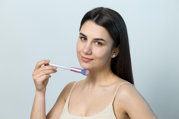 Nahaufnahme eines jungen Mädchens in einem hellen Oberteil auf weißem Hintergrund, das ein Gesichts-Make-up macht Eine hübsche Frau hält einen Kosmetikpinsel in der Nähe ihres Gesichts und lächelt