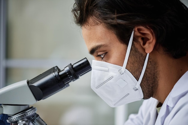 Nahaufnahme eines jungen Mannes mit Atemschutzmaske, der im Labor unter dem Mikroskop schaut