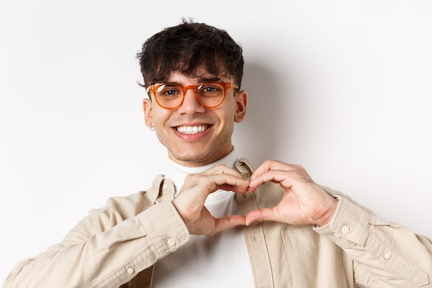 Nahaufnahme eines jungen natürlichen Kerls mit Brille, der lächelt, Herzliebe zeigt, Geste, stehend auf weißem Hintergrund