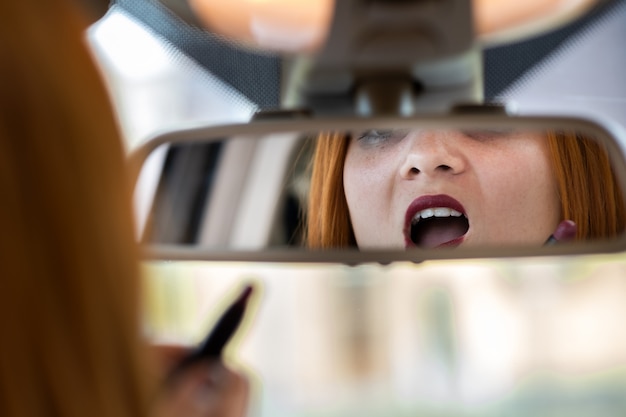 Nahaufnahme eines jungen rothaarigen Frauenfahrers, der ihr Make-up mit dem dunkelroten Lippenstift korrigiert, der im Auto-Rückspiegel hinter dem Lenkrad eines Fahrzeugs schaut.