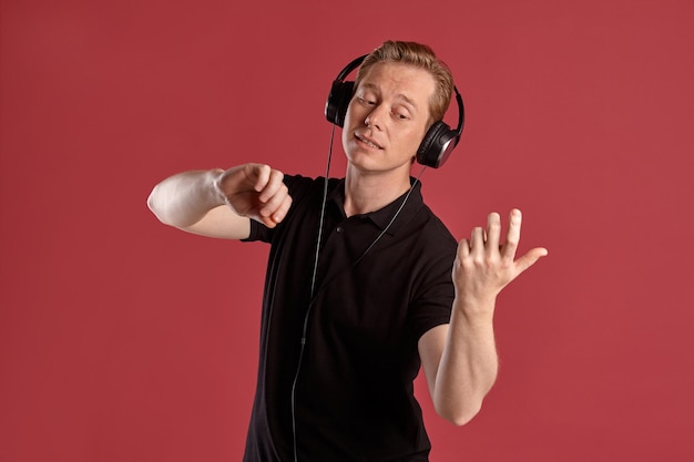 Nahaufnahme eines jungen, stylischen Ginger-Mannes in einem stylischen schwarzen T-Shirt, der Musik über Kopfhörer hört und so tut, als würde er Geige spielen, während er auf rosafarbenem Studiohintergrund posiert