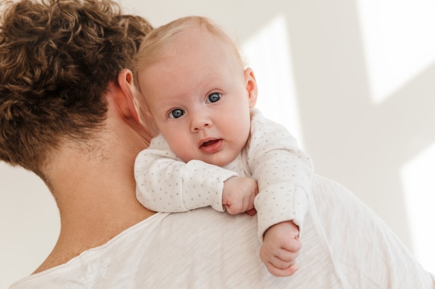 Nahaufnahme eines jungen Vaters, der seinen kleinen Sohn hält, während er drinnen steht, Baby schaut direkt