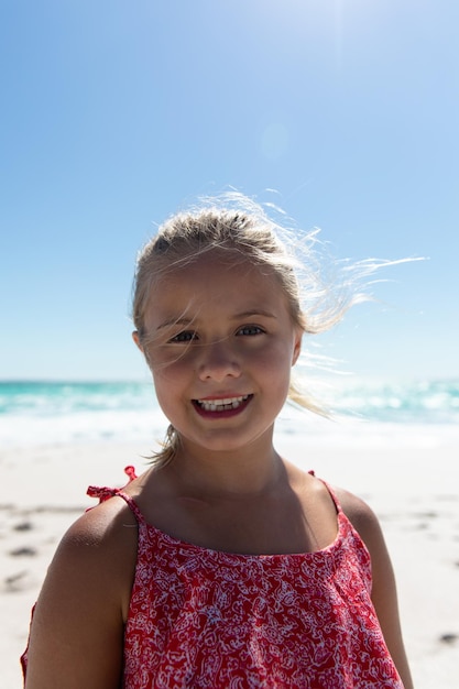 Nahaufnahme eines kaukasischen Mädchens, das am Strand mit blauem Himmel und Meer im Hintergrund steht und zur Kamera lächelt