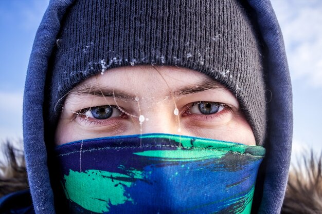Nahaufnahme eines kaukasischen Mannes Gesicht mit Schal und Kapuzen-Winterjacke mit Frost auf den Augenbrauen in die Kamera schaut