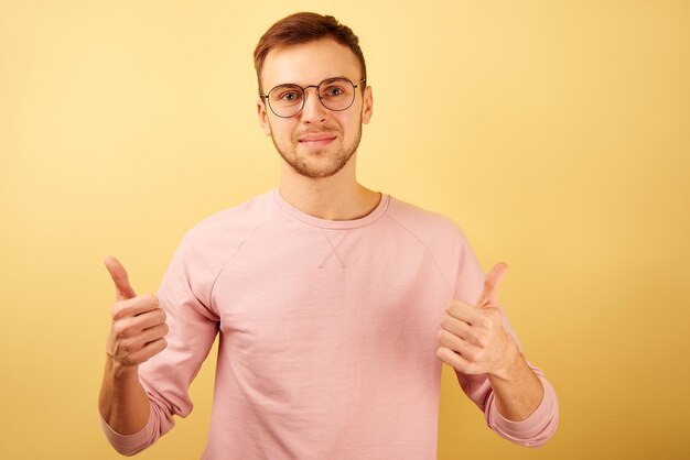 Nahaufnahme eines kaukasischen Mannes mit Brille und lächelndem Gesicht, der Daumen nach oben zeigt, isoliert auf gelbem Hintergrund in rosafarbener Freizeitkleidung