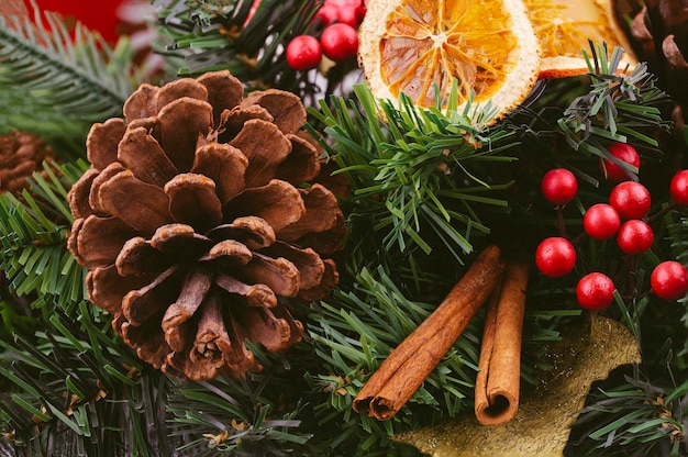 Foto nahaufnahme eines kegels auf einem tannenzweig, dekoriert mit zimtstangen, roten beeren, getrockneten orangen, weihnachtshintergrund