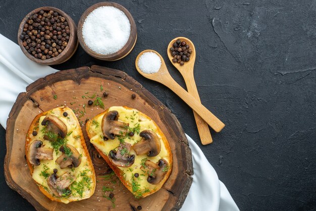 Nahaufnahme eines köstlichen Snacks mit Pilzen auf Holzbrett und Gewürzen auf weißem Handtuch auf schwarzem Hintergrund