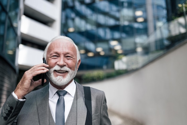 Nahaufnahme eines lächelnden älteren Geschäftsmannes, der am Handy gegen das Gebäude diskutiert