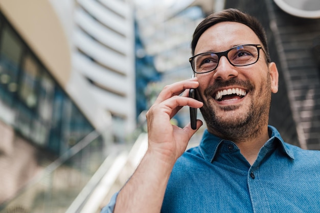 Nahaufnahme eines lächelnden Unternehmers, der mit seinem Smartphone vor einem Bürogebäude spricht