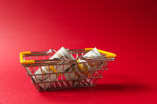 Nahaufnahme eines Lebensmittelkorbs aus Metall zum Einkaufen im Supermarkt mit abgesenkten gelben Griffen mit Dollarbündeln einzeln auf rotem Hintergrund. Konzept des Einkaufens. Kopieren Sie Platz für Werbung.