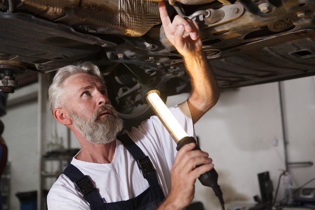 Nahaufnahme eines leitenden Autoservice-Mitarbeiters, der eine Lampe hält und das Auto auf dem Autolift untersucht?