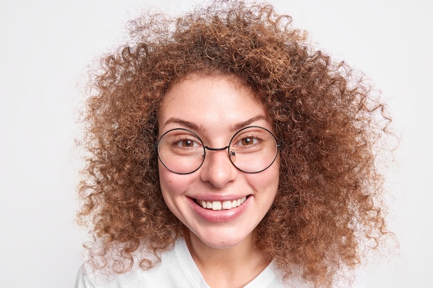 Nahaufnahme eines lockigen, lächelnden europäischen weiblichen Modells mit natürlichem Make-up hat glücklichen Ausdruck, perfekte Zähne stehen entspannt im Innenbereich und tragen runde Brillen isoliert über weißer Wand.