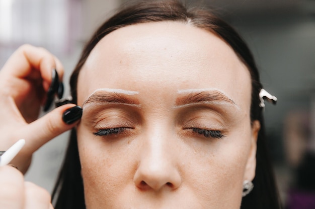 Foto nahaufnahme eines mädchengesichts der meister hält einen weißen bleistift und zeichnet die form der augenbrauen für dauerhaftes make-up