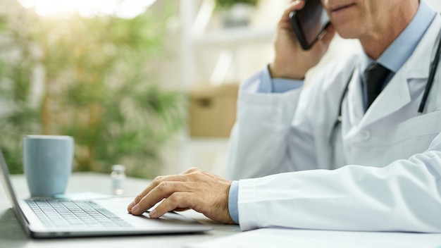 Nahaufnahme eines männlichen Arztes, der am Tisch sitzt und am Notebook arbeitet, während er in der Klinik telefoniert