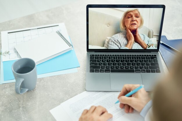 Nahaufnahme eines männlichen Arztes, der mit Notebook am Tisch sitzt und Notizen macht, während die alte Dame den Hals berührt und die Symptome beschreibt