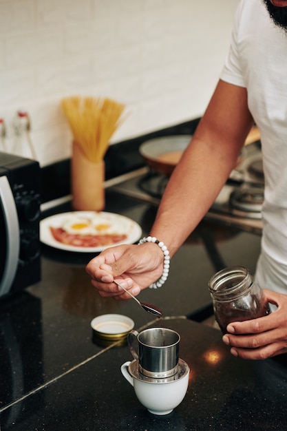 Nahaufnahme eines Mannes, der einen Löffel gemahlenen Kaffeepulvers in einen Aluminiumtropfer setzt, wenn er zu Hause Kaffee im vietnamesischen Stil zubereitet