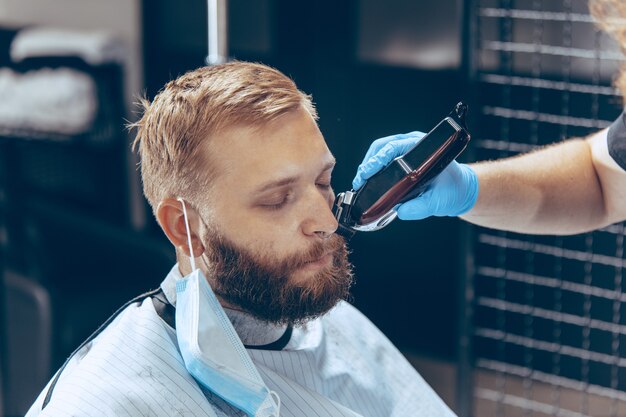 Nahaufnahme eines Mannes, der sich während der Coronavirus-Pandemie im Friseursalon die Haare schneiden lässt und eine Maske trägt. Professioneller Friseur mit Handschuhen. Covid-19, Schönheits-, Selbstpflege-, Stil-, Gesundheits- und Medizinkonzept.