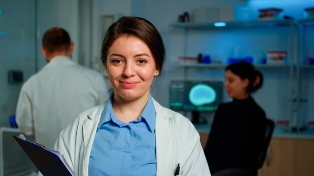 Nahaufnahme eines medizinischen Neurologen-Forschers, der in die Kamera lächelt, die im Labor steht und Clipdoard hält, während ein Mitarbeiter mit dem Patienten über die Gesundheitsversorgung des Gehirns diskutiert