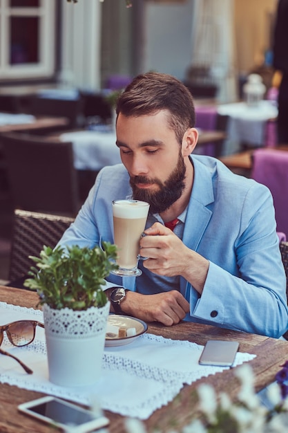 Nahaufnahme eines modischen bärtigen Mannes mit stilvollem Haarschnitt, trinkt ein Glas Cappuccino und sitzt in einem Café im Freien.