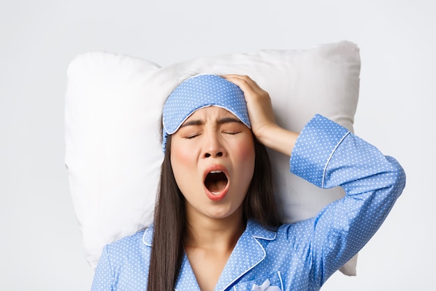 Nahaufnahme eines müden und schläfrigen süßen asiatischen Mädchens in Schlafmaske und Pyjama, Gähnen und Berühren des Kopfes als Kopfschmerzen nach einer Kater-Übernachtungsparty, Schlaflosigkeitsprobleme, auf Kissen liegend.
