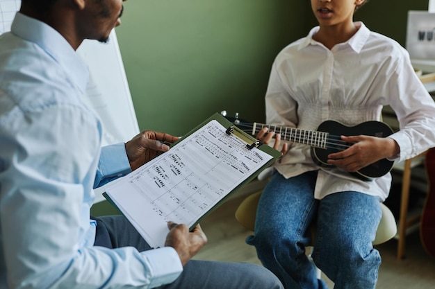 Nahaufnahme eines Musiklehrers mit Musiknoten, der mit einem Schulmädchen diskutiert, das Ukulele spielt und