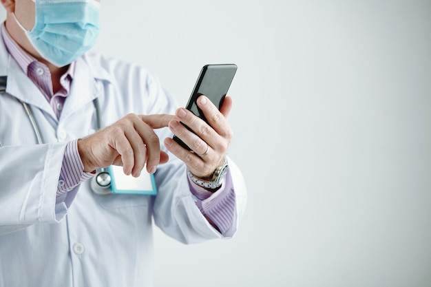 Nahaufnahme eines nicht erkennbaren Arztes in Maske und weißem Kittel, der die Nachricht am Telefon vor grauem Hintergrund überprüft