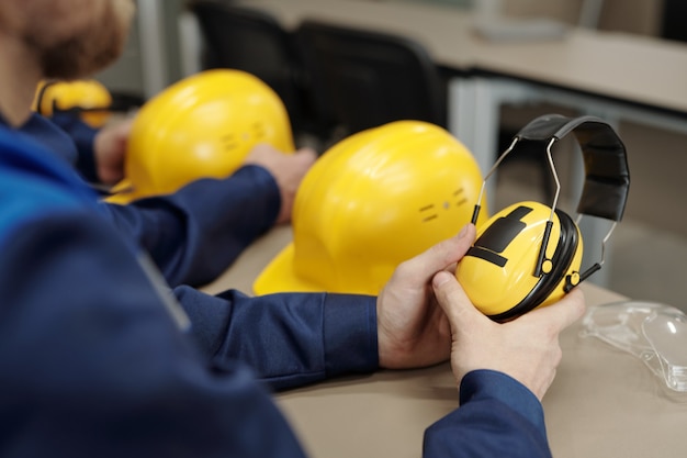 Nahaufnahme eines nicht erkennbaren Fabrikarbeiters, der mit Schutzhelm und Schutzbrille am Tisch sitzt und schalldichte Schutzkopfhörer hält