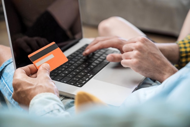 Nahaufnahme eines nicht erkennbaren schwulen Paares, das einen Laptop benutzt, während es zu Hause auf einer Couch sitzt und mit einer Kreditkarte online einkauft