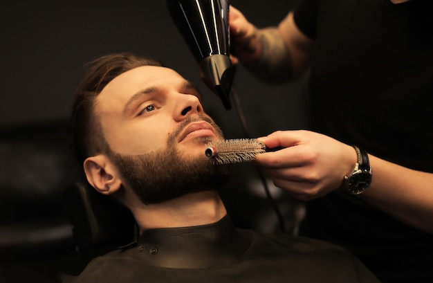Nahaufnahme eines professionellen Friseurs, der den Bart seines jungen Kunden pflegt, der während des Eingriffs in den Spiegel schaut.