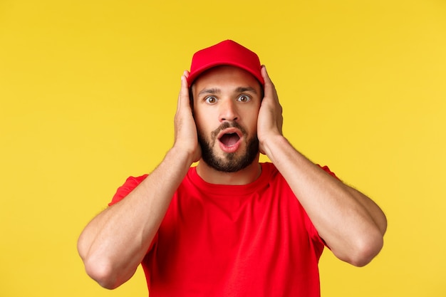 Nahaufnahme eines schockierten, keuchenden männlichen Kurierangestellten in roter Uniformmütze und T-Shirt bedecken die Ohren und starren ...