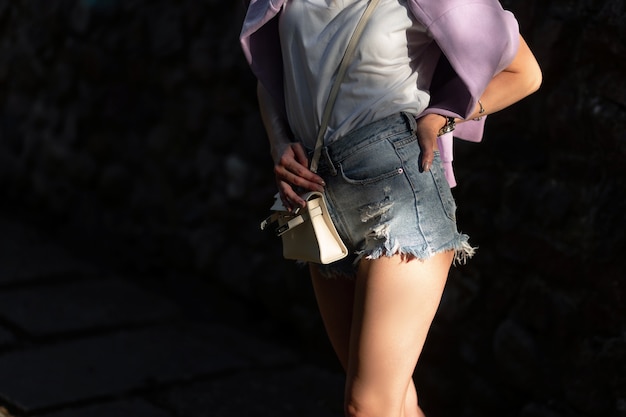 Nahaufnahme eines schönen weiblichen Körpers in modischen Jeansshorts mit weißem T-Shirt mit Handtasche im Freien auf dunklem Hintergrund