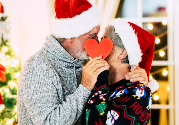 Nahaufnahme eines Seniorenpaares, das einen Weihnachtsmannshut trägt und zu Hause in Herzform steht